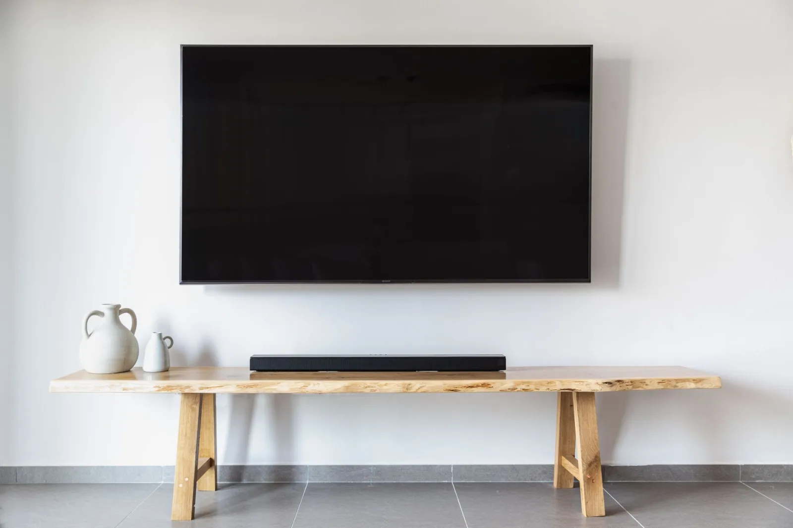 Duży telewizor zamontowany na białej ścianie nad drewnianą ławą z soundbarem i dzbanami.