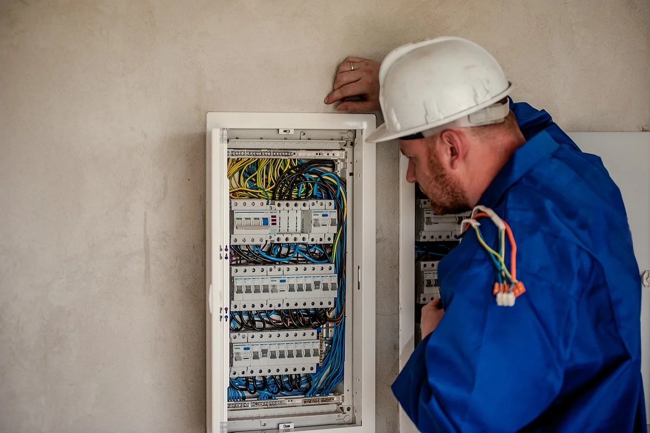 Elektryk w białym kasku i niebieskim ubraniu sprawdza rozdzielnicę z modułowymi wyłącznikami na szynie DIN.