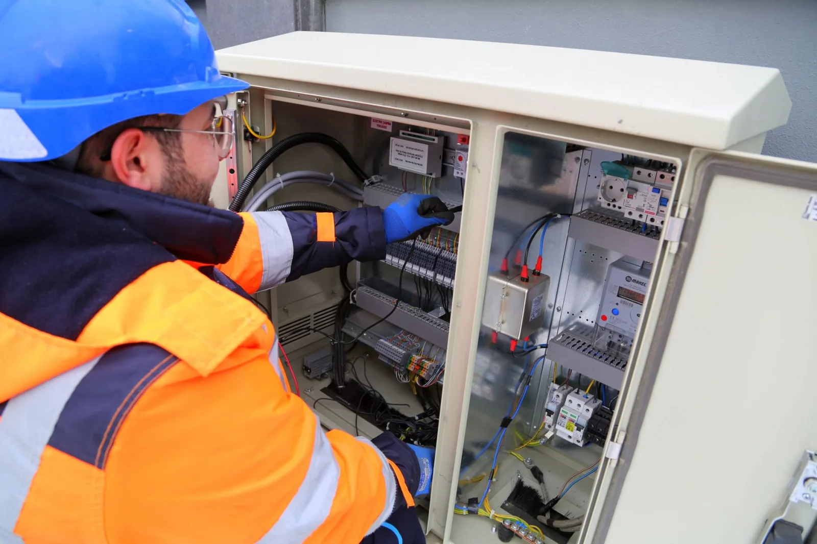 Elektryk w odblaskowej kurtce i kasku sprawdza otwartą skrzynkę elektryczną na zewnątrz budynku